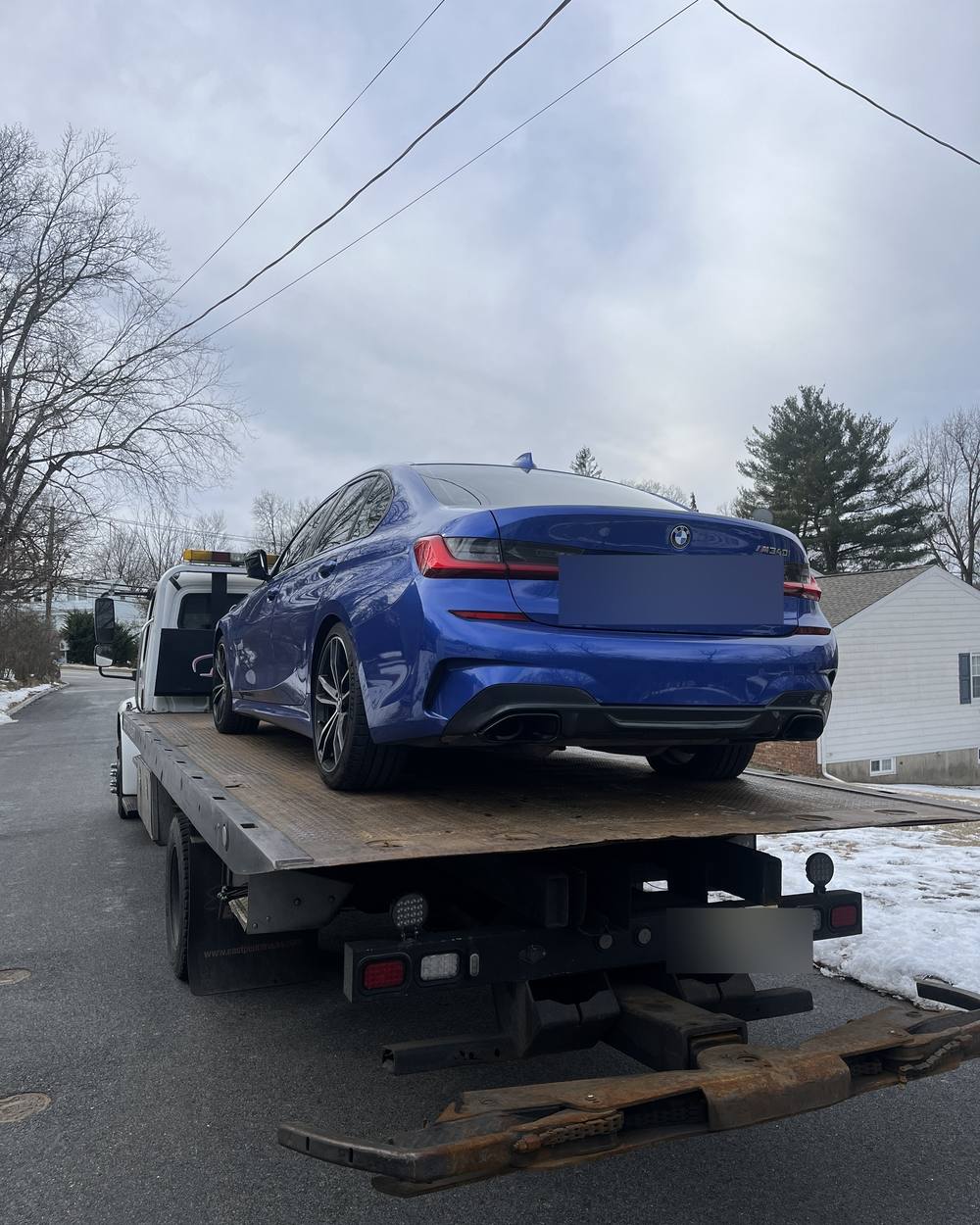 A blue BMW M340i strapped down on a flatbed tow truck on a snowy North Jersey residential street.