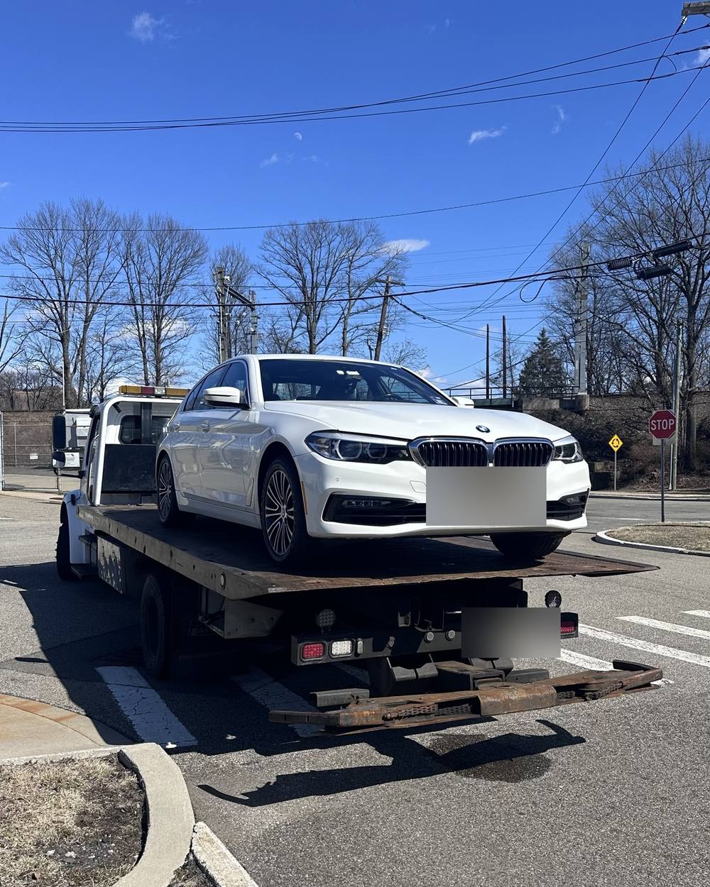 A white BMW 5 Series loaded on a flatbed tow truck at an intersection in North Jersey.