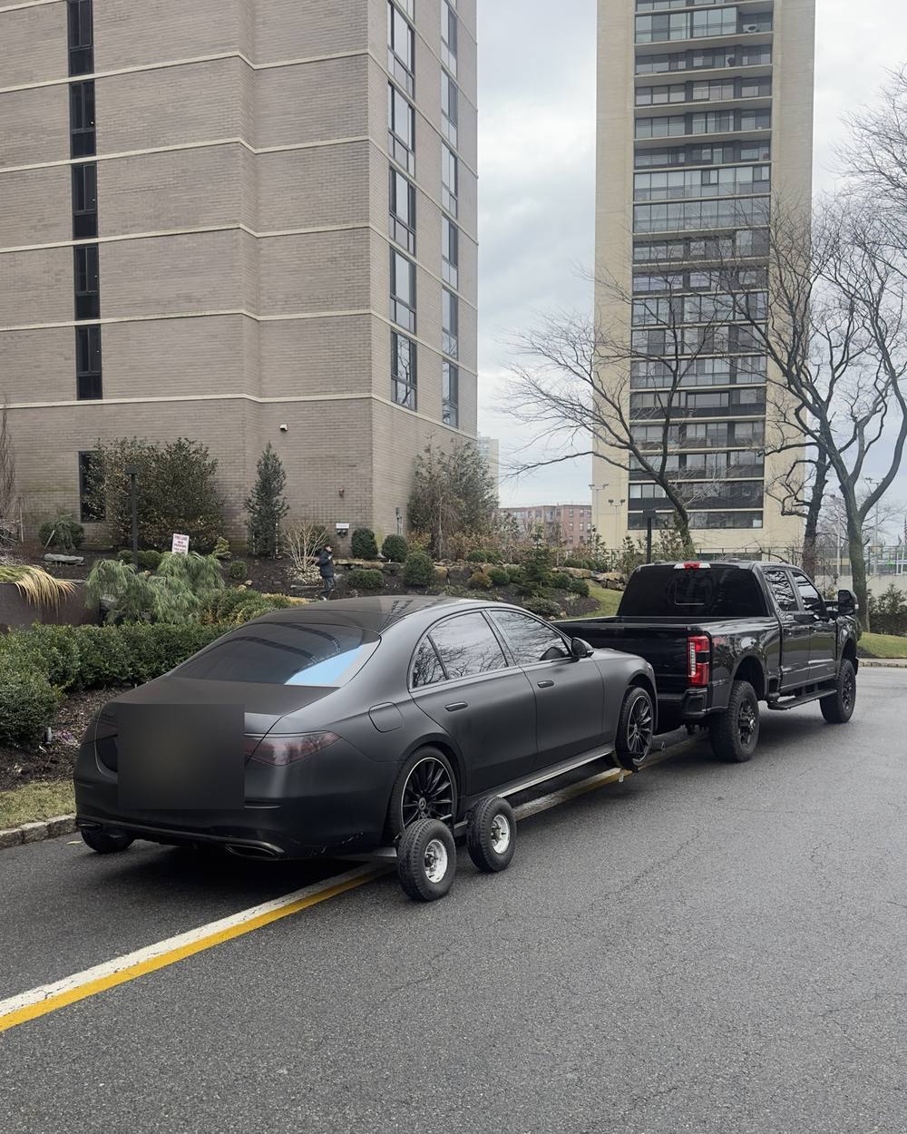 A matte black Mercedes-Benz S-Class raised on tow dollies behind a pickup, ready to move from a residential high-rise driveway.